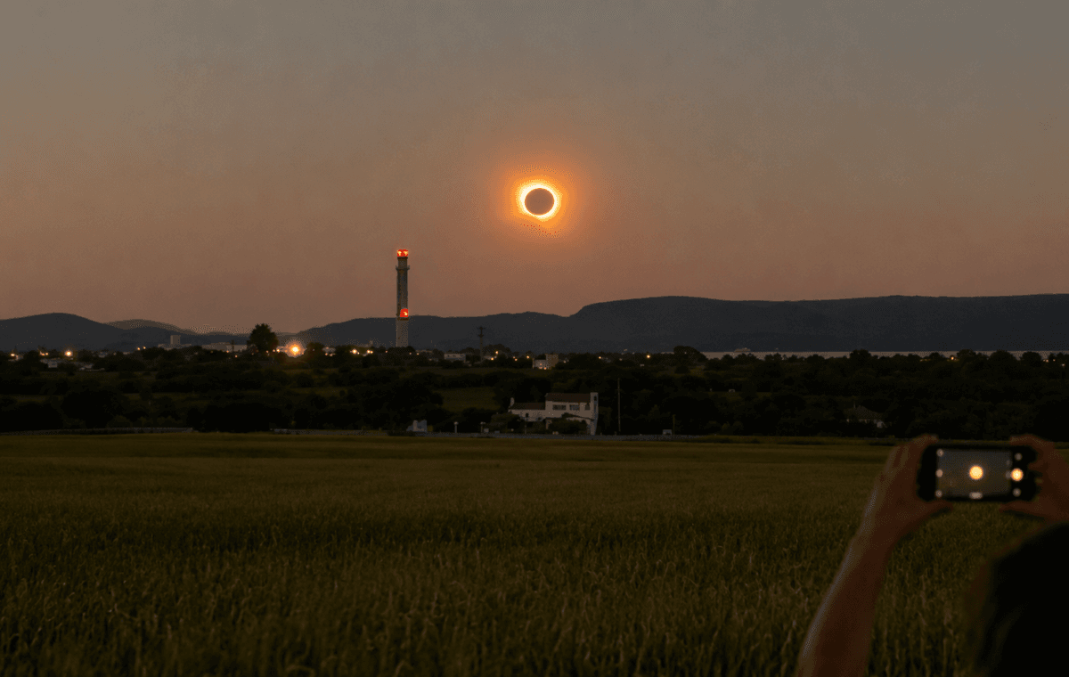 Ilustração por IA mostra eclipse solar próximo ao horizonte enquanto uma pessoa registra o fenômeno.