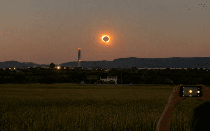 Ilustração por IA mostra eclipse solar próximo ao horizonte enquanto uma pessoa registra o fenômeno.