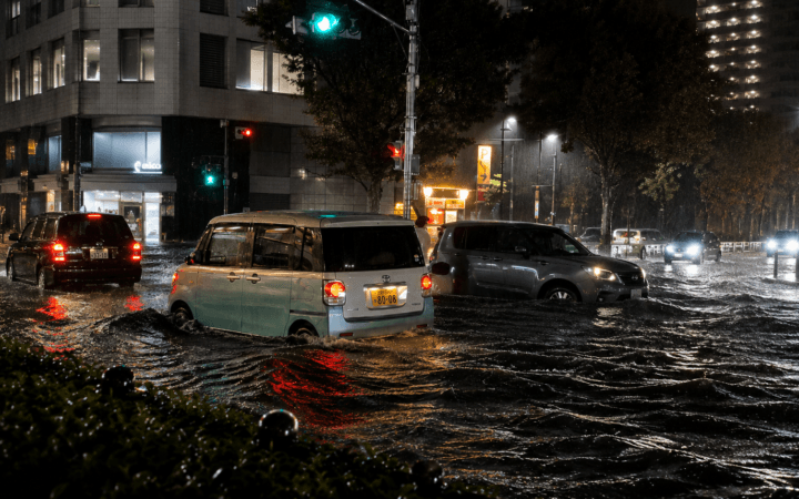 Ilustração por IA mostra veículos atravessando rua alagada durante chuva intensa em uma área urbana do Japão.